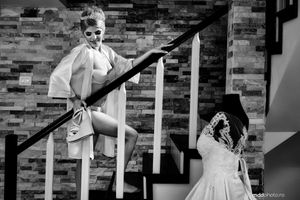 A black and white artistic wedding photo of someone in sunglasses on stairs while someone in a dress kneels below.