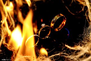 Two golden wedding rings surrounded by flames and cobwebs in a dark, moody setting.