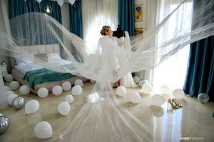 Flowing white veil billows dramatically in a room decorated with white balloons and blue curtains on a sunny morning.