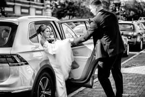 A black and white photo shows a bride getting assistance exiting a car on what appears to be their wedding day.