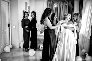 Bridesmaids in black dresses help the bride get ready in a room with white balloons and curtains.