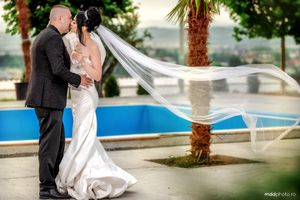 A couple in formal wedding attire embrace beside a blue swimming pool with a flowing white veil catching the breeze.