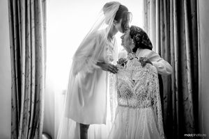 A bridal preparation scene in black and white showing a wedding dress on display while someone adjusts it near a window.