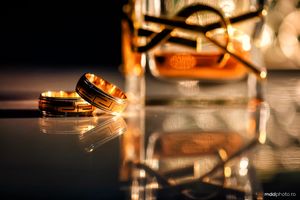 Two golden wedding rings reflecting on a shiny surface with warm bokeh lights in the background.