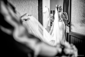 Black and white wedding photograph showing a bride in an elegant white dress with lace details standing in a doorway.