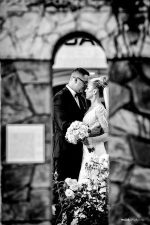 A romantic black and white wedding photo captured under a stone archway with a bouquet of white flowers.