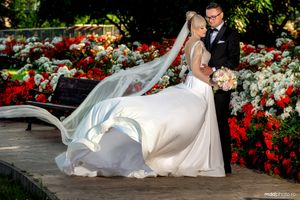 A newlywed couple poses in a garden with vibrant red flowers as the bride's white dress flows in the wind.