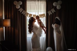 A silhouetted figure lifts a child by window light with wedding decorations and a dress hanging in a dark room.