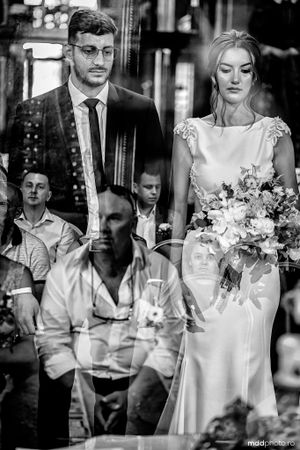 Black and white wedding photo showing couple with artistic reflections in window glass and floral bouquet.