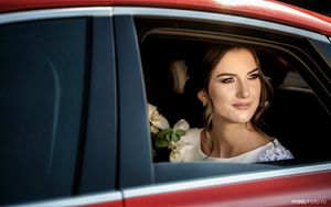 A bride sits in the back of a red car on her wedding day, smiling through the window with elegant makeup and hair.