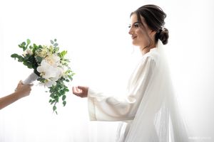 Someone in a white wedding dress receives a bouquet of white roses and greenery against a light background.