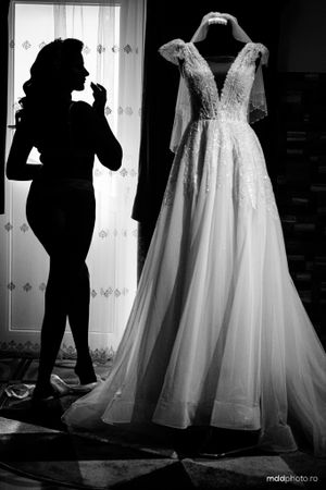 Black and white photo of an elegant wedding dress on display with a silhouette figure standing near a window.