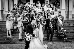 Black and white wedding photo shows wedding party gathered on church steps as newlyweds share romantic moment.