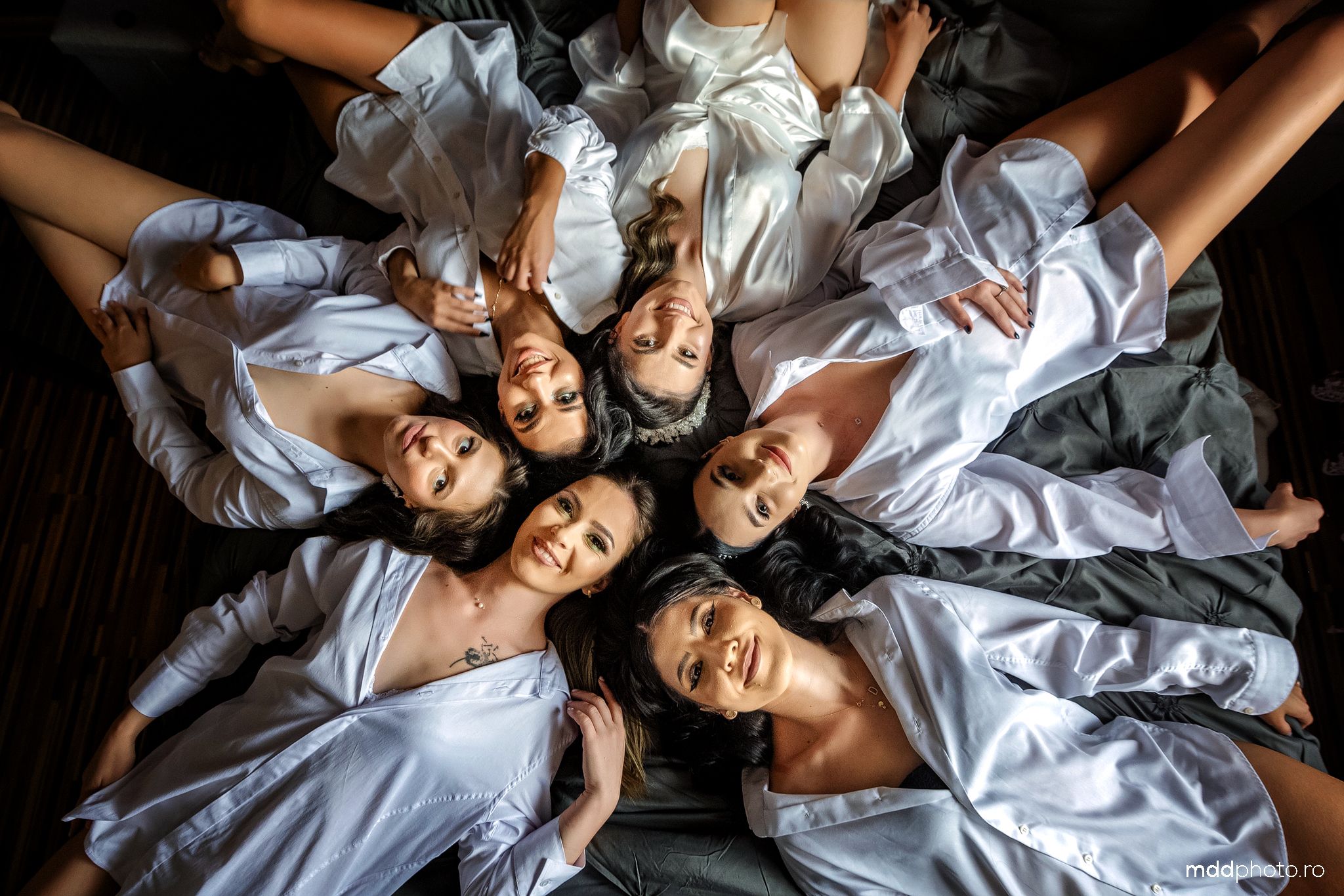 People wearing white button-down shirts laying in a circle formation on a dark floor looking up at the camera.