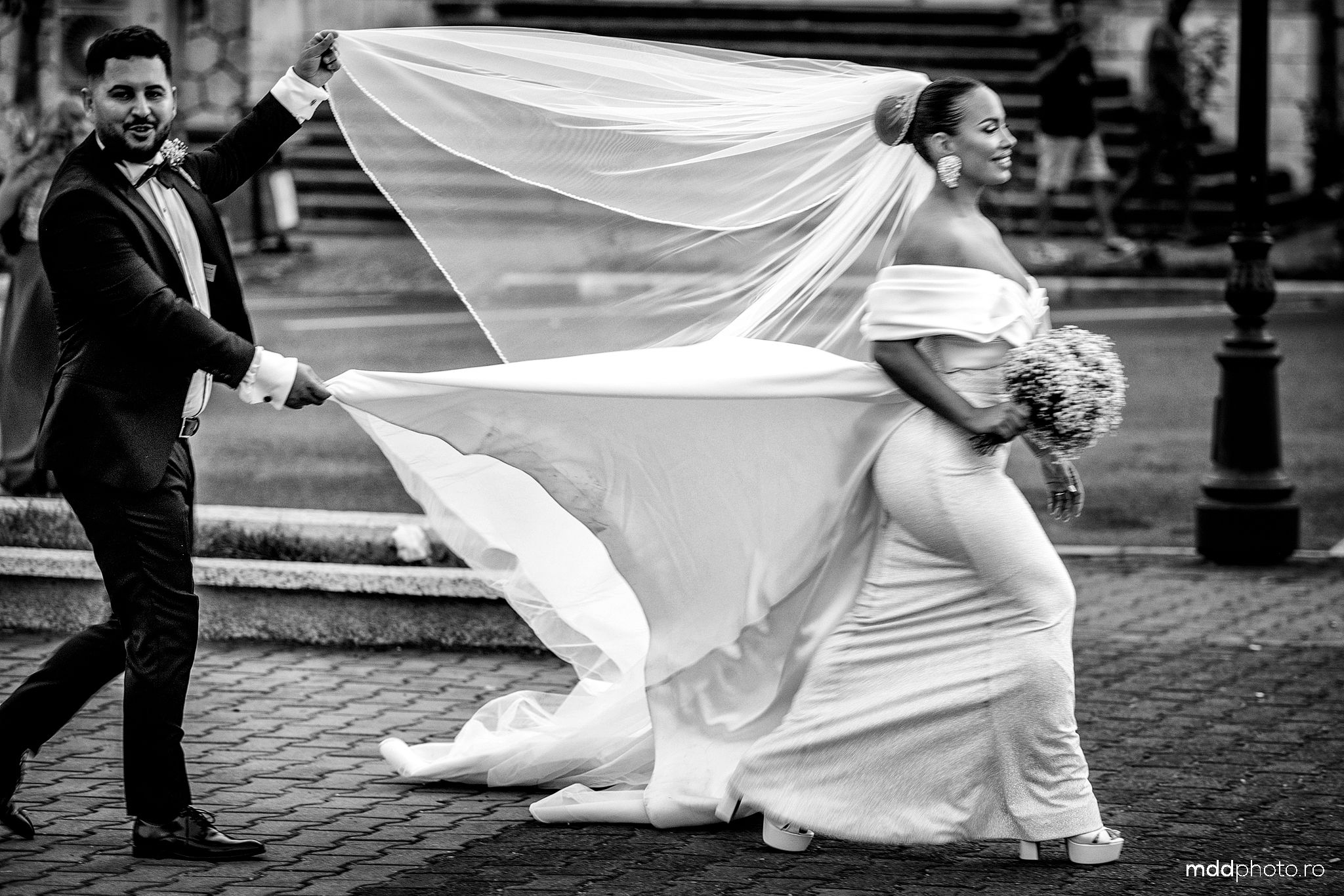 A dramatic black and white wedding photograph shows a flowing veil caught in the wind on a city street.