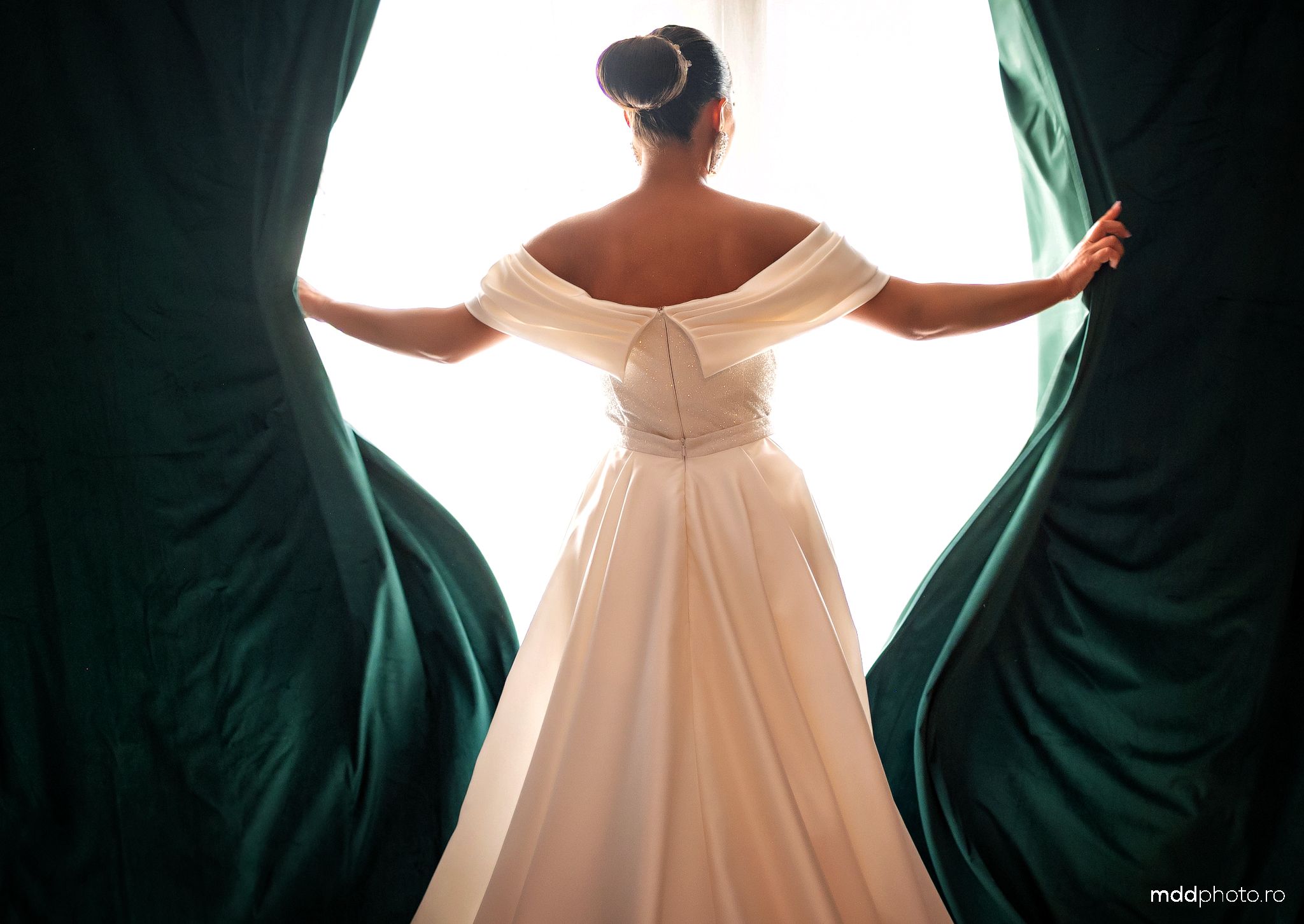 Back view of a bride in an off-shoulder white gown parting emerald green curtains in dramatic lighting.