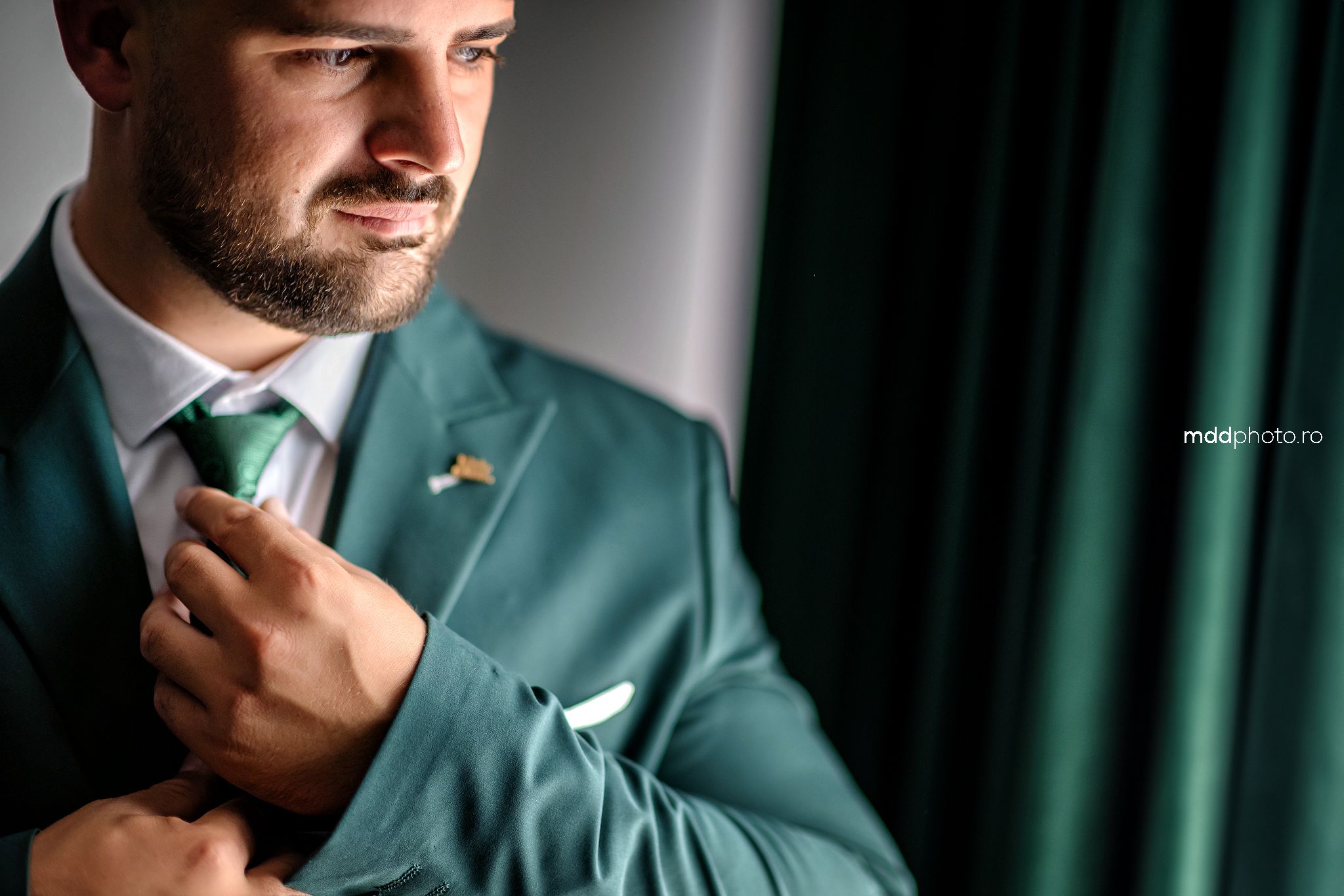 A groom in a mint green suit adjusts his tie while standing near emerald curtains in dramatic lighting.