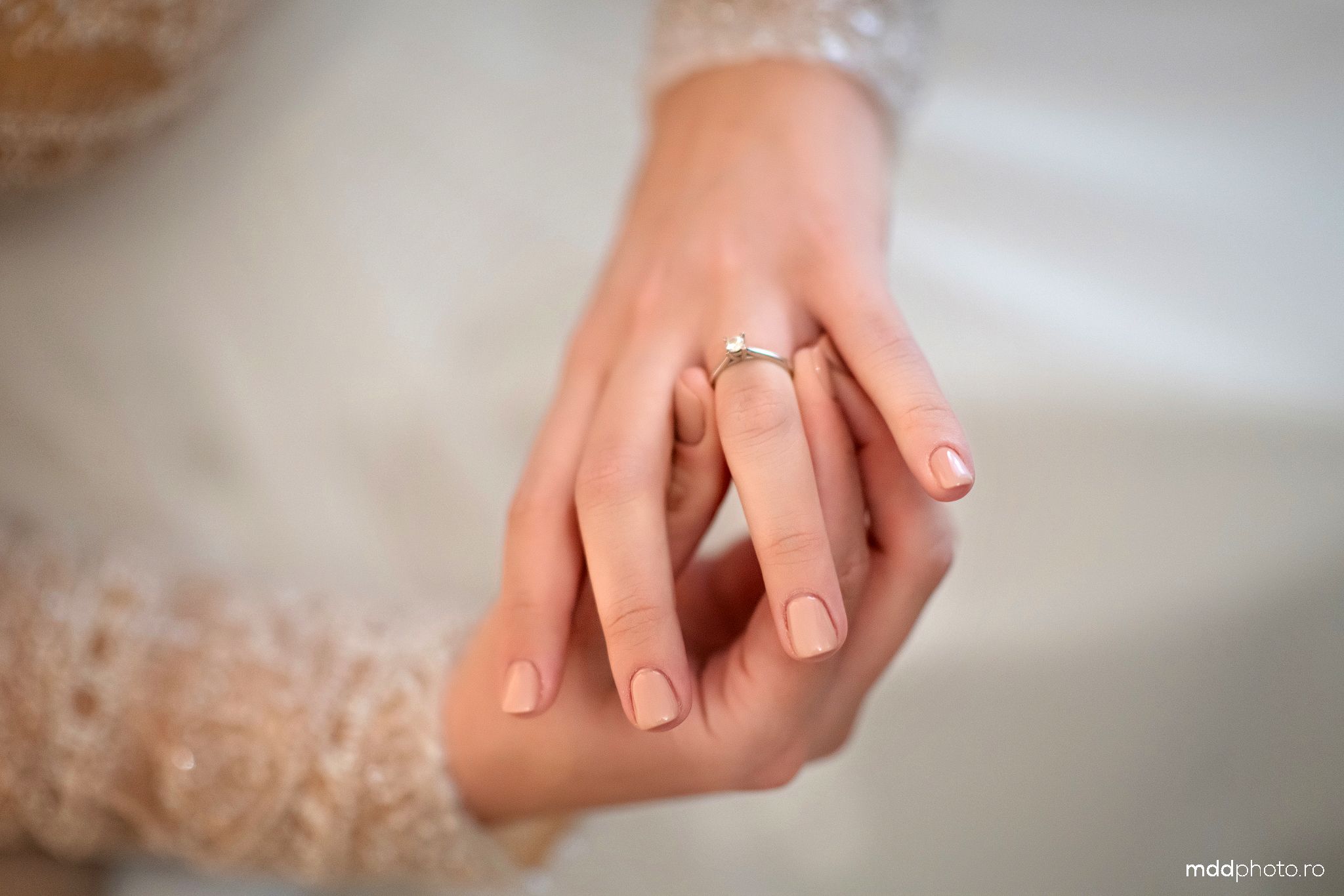 Close up of a delicate engagement ring on a woman's hand with light pink manicured nails and lace details.