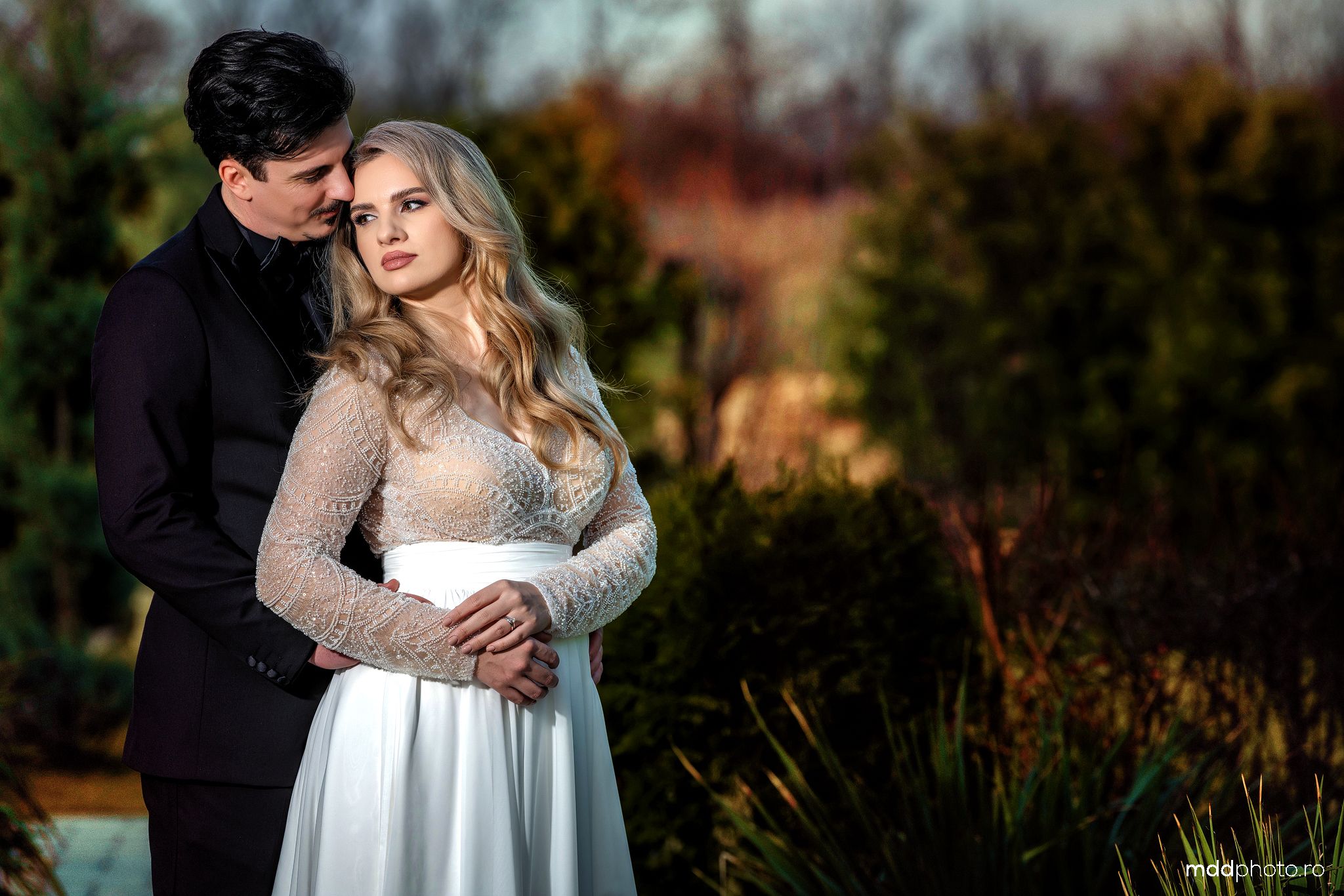 A romantic couple embraces outdoors in formal attire with natural foliage in the background during golden hour.