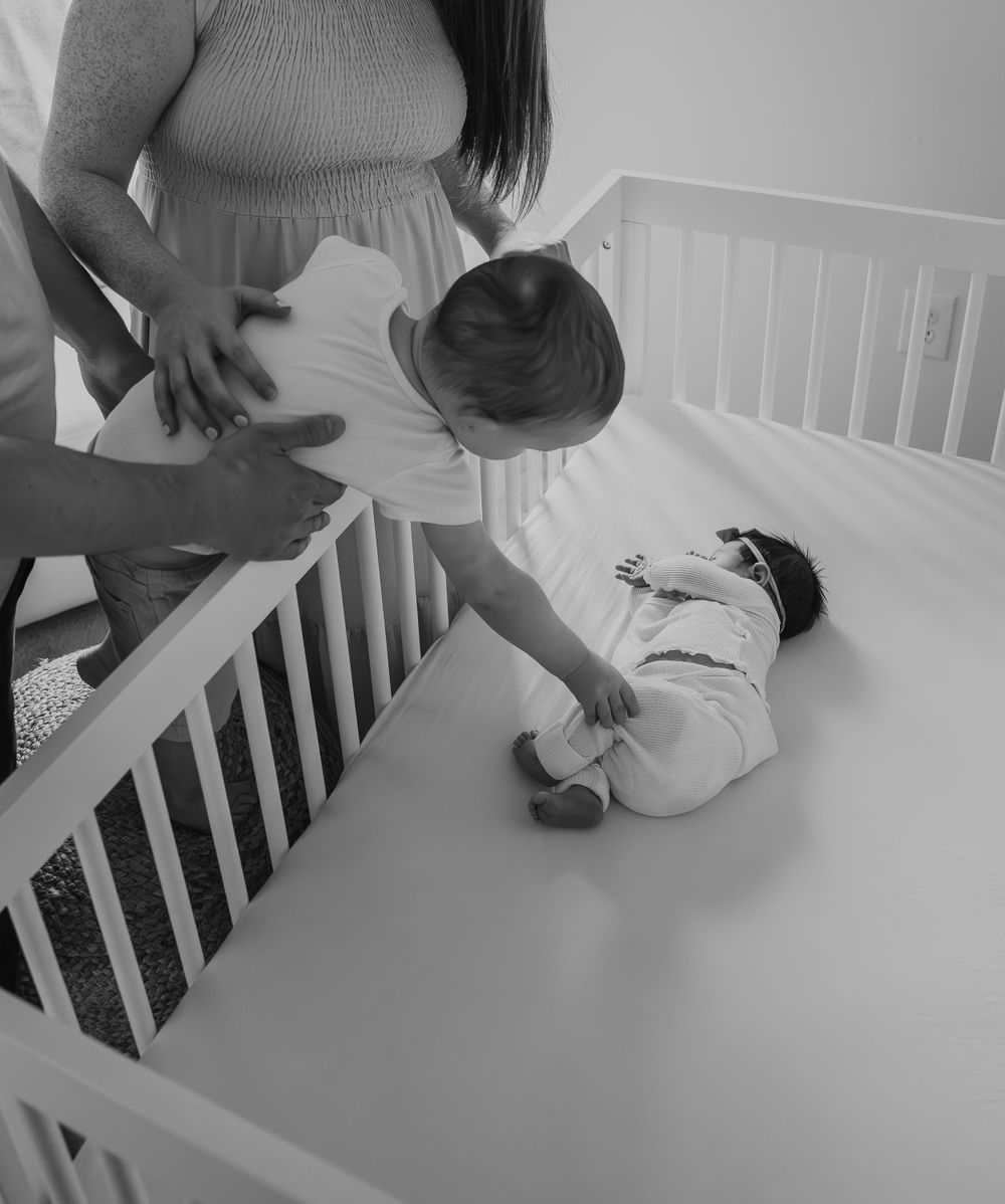 Overhead black and white photo of toddler reaching into crib to touch newborn sibling.