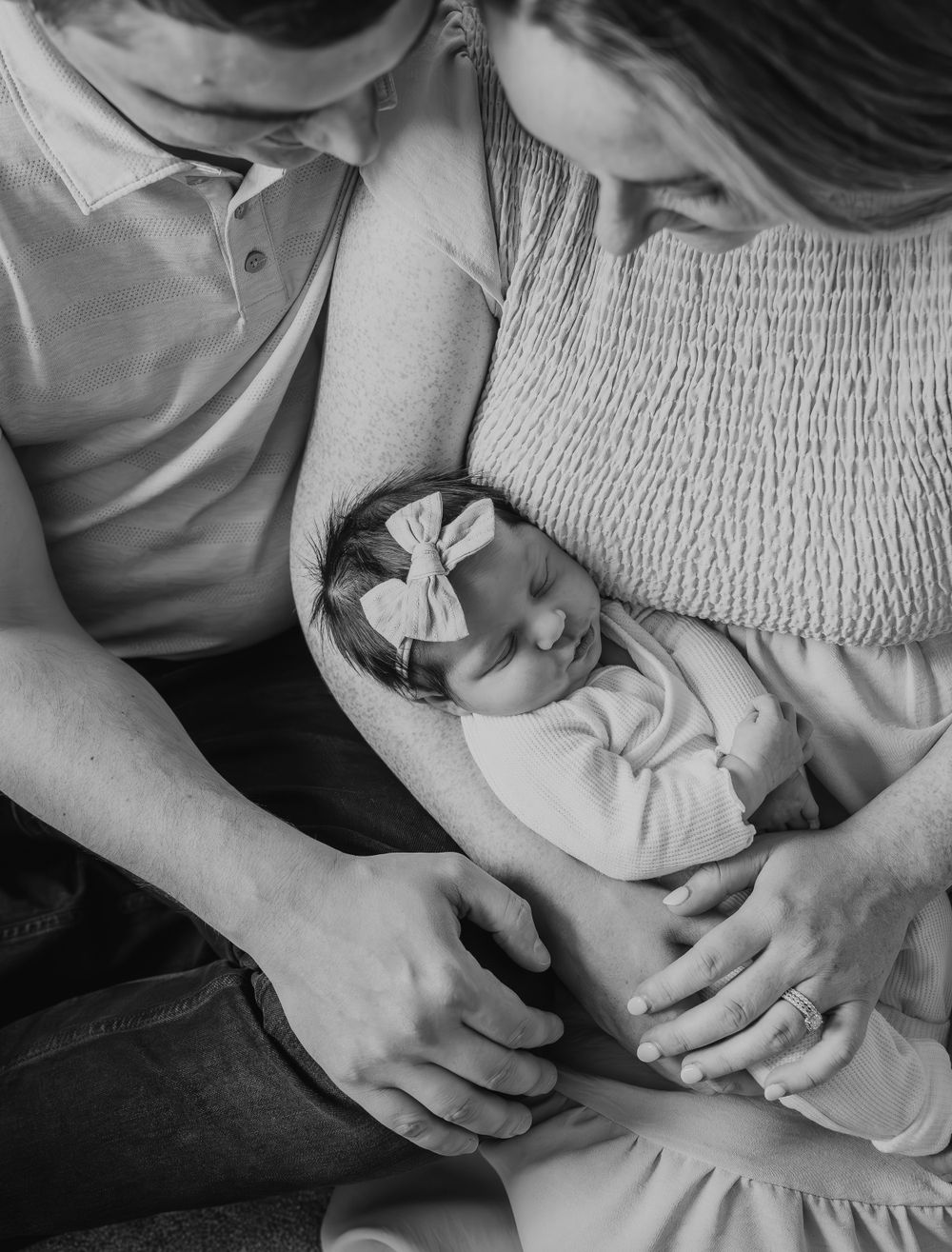 Black and white close-up of parents cradling sleeping newborn baby girl with bow headband.