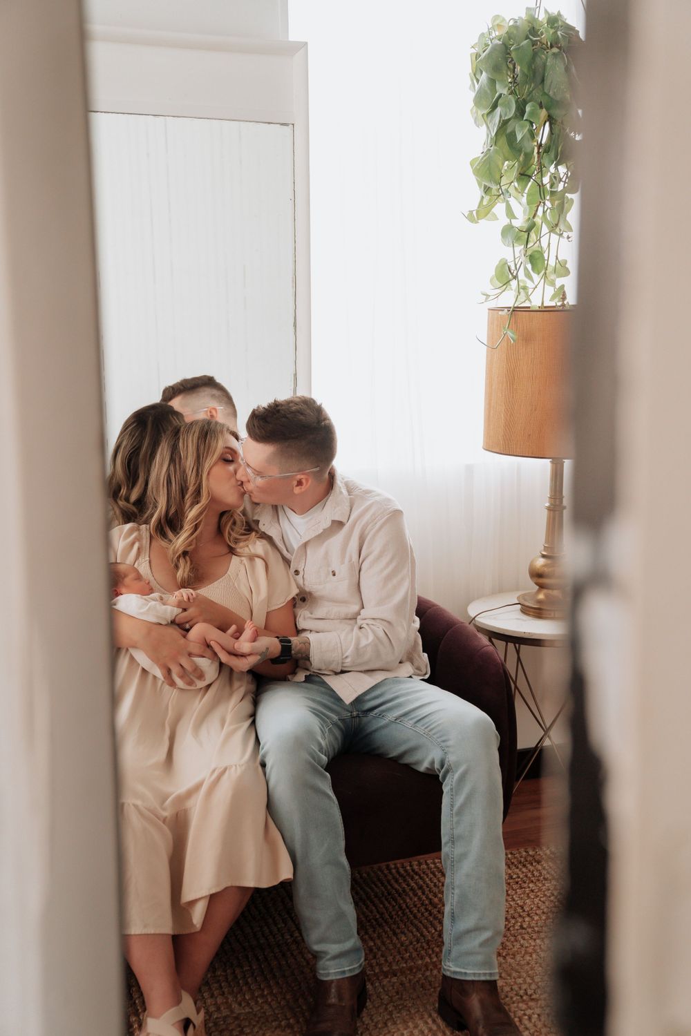 New parents sharing a kiss while holding their newborn baby together on a couch in a cozy room.