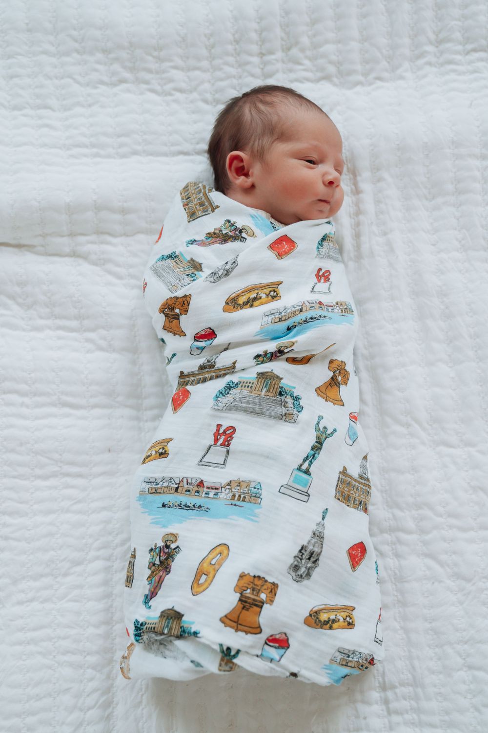 Peaceful newborn baby swaddled in a Philadelphia landmarks themed muslin blanket on white bedding.