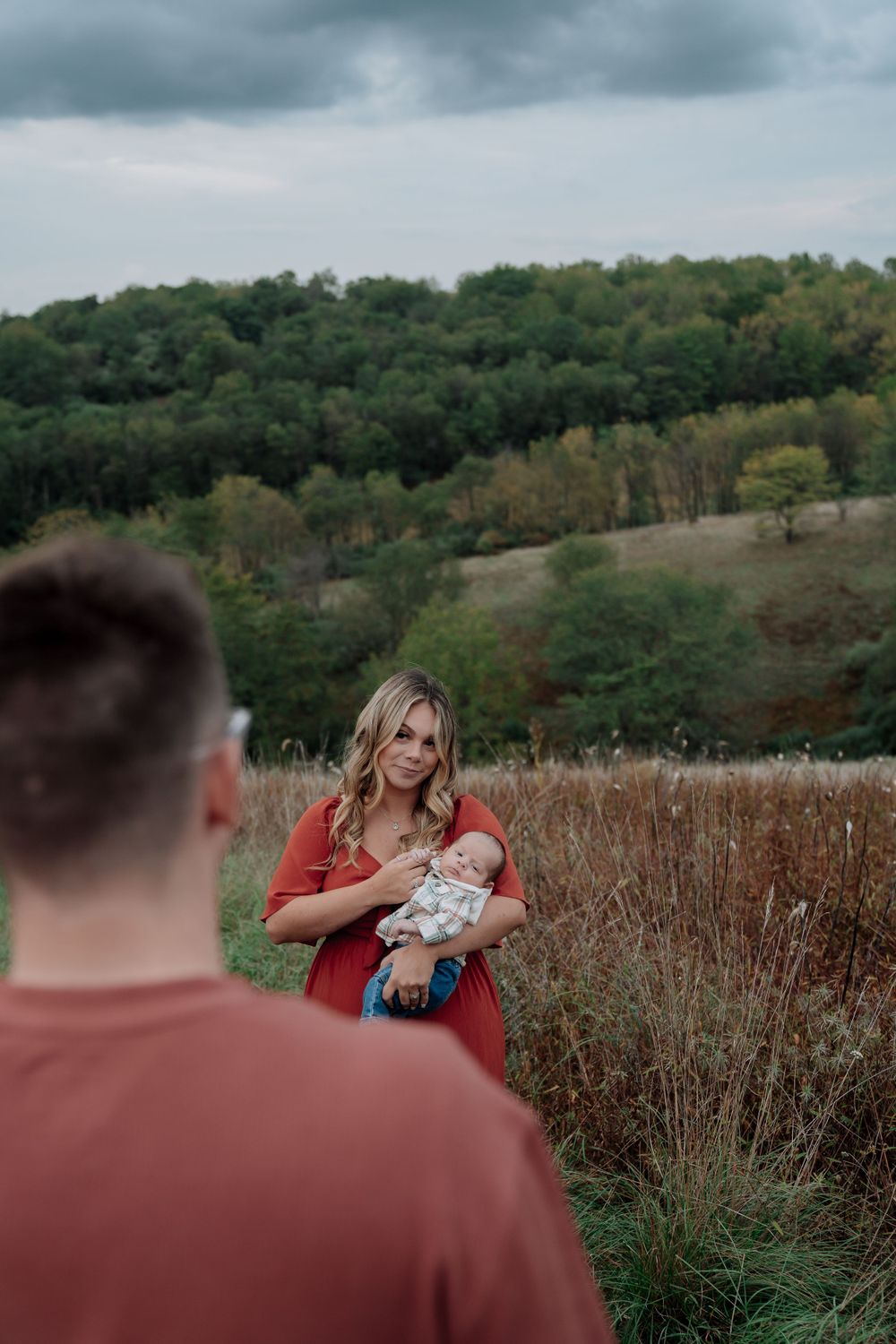 Woman in red dress holding newborn baby in a scenic outdoor field setting.