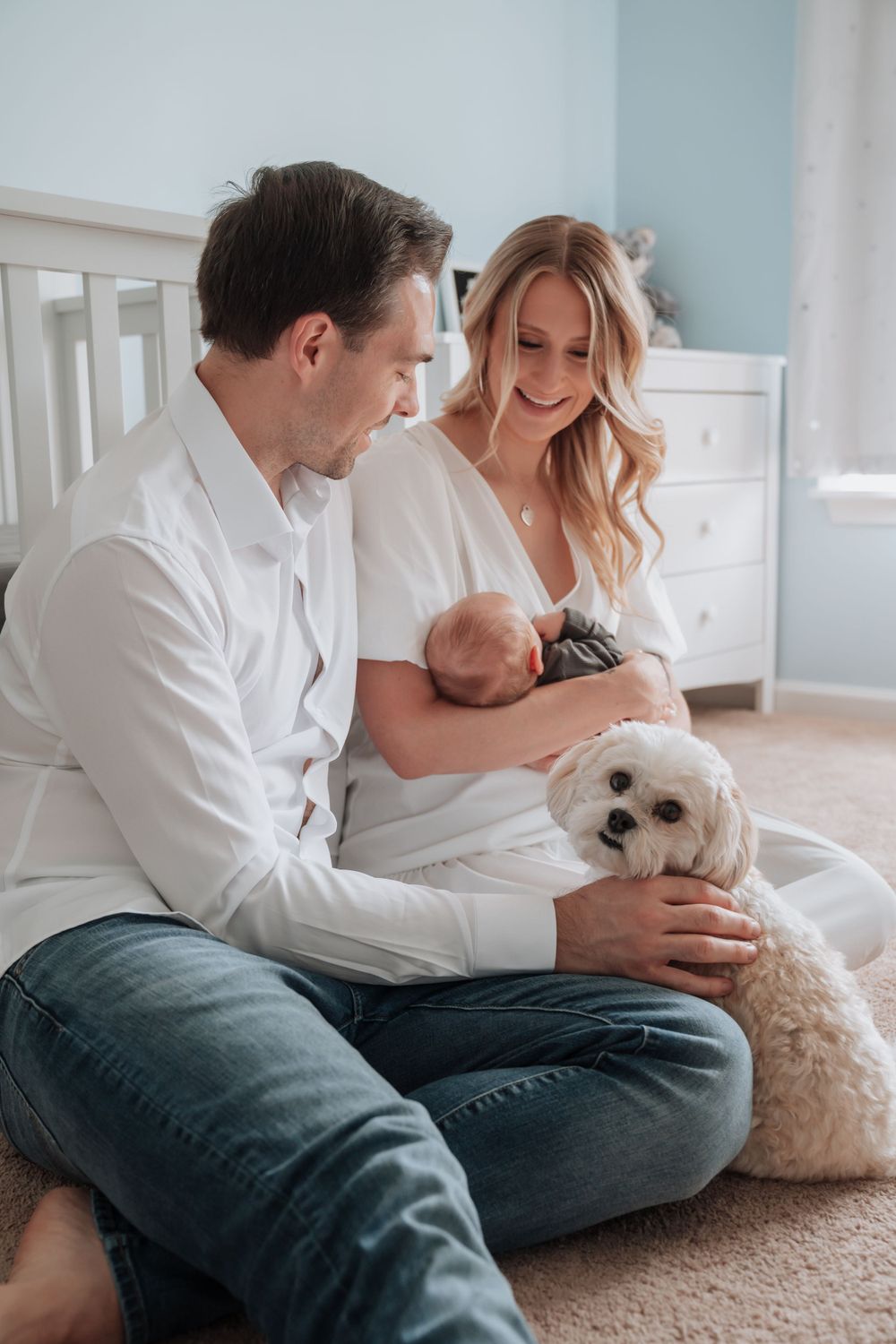 Young family sits on nursery floor holding newborn baby with white fluffy dog.