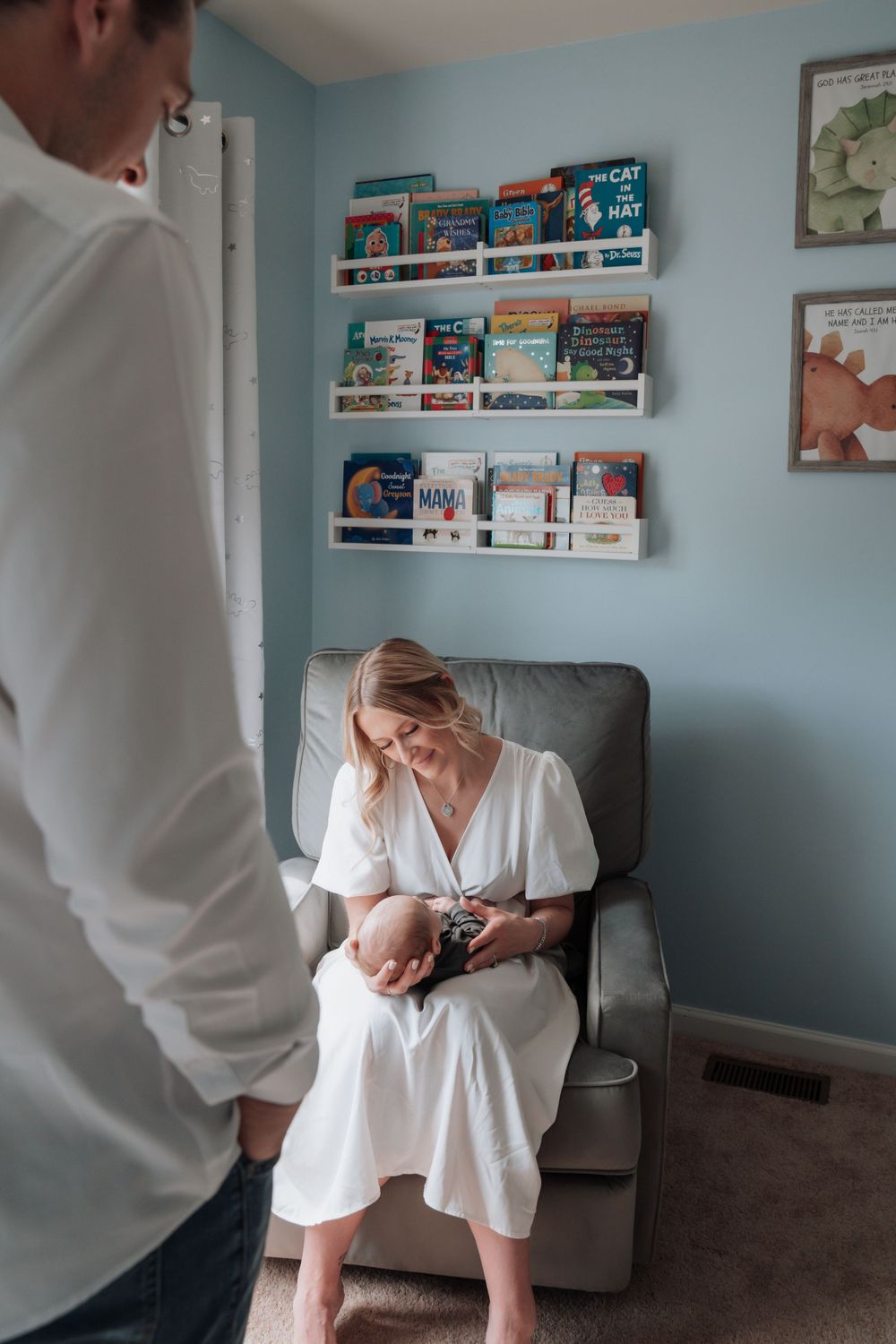 Mother in white dress sits in nursery glider smiling at newborn while father watches.
