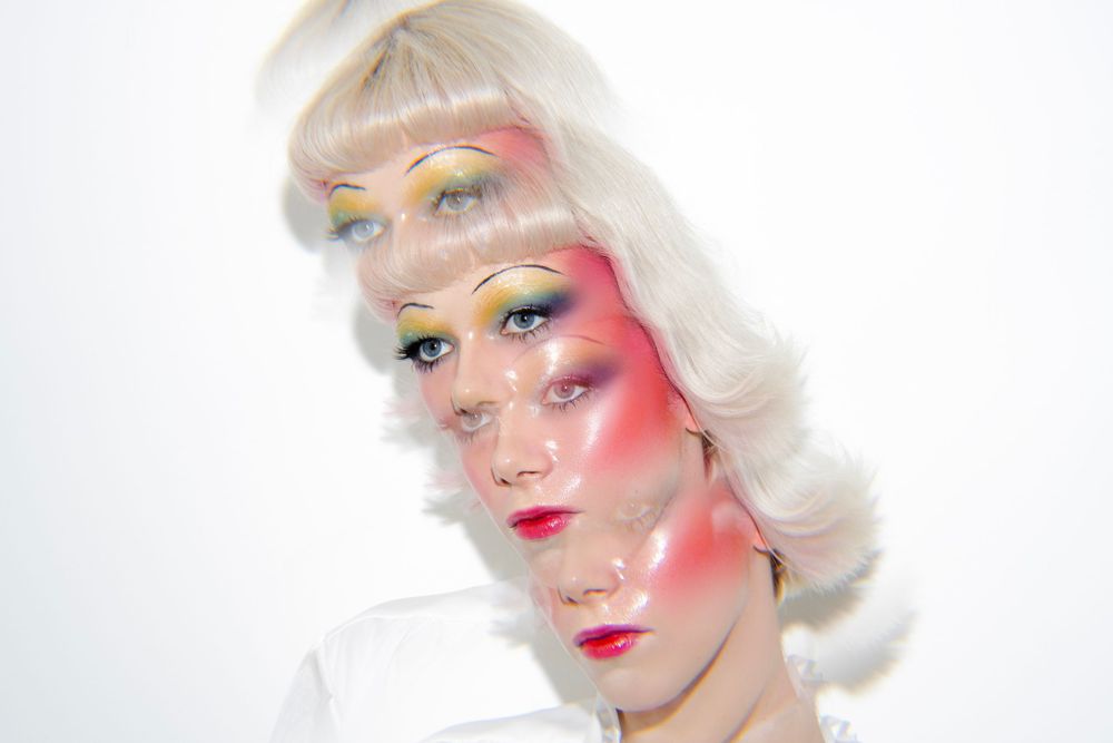 Creative beauty portrait with artistic pink and orange makeup, platinum blonde hair, and ethereal lighting against white background.