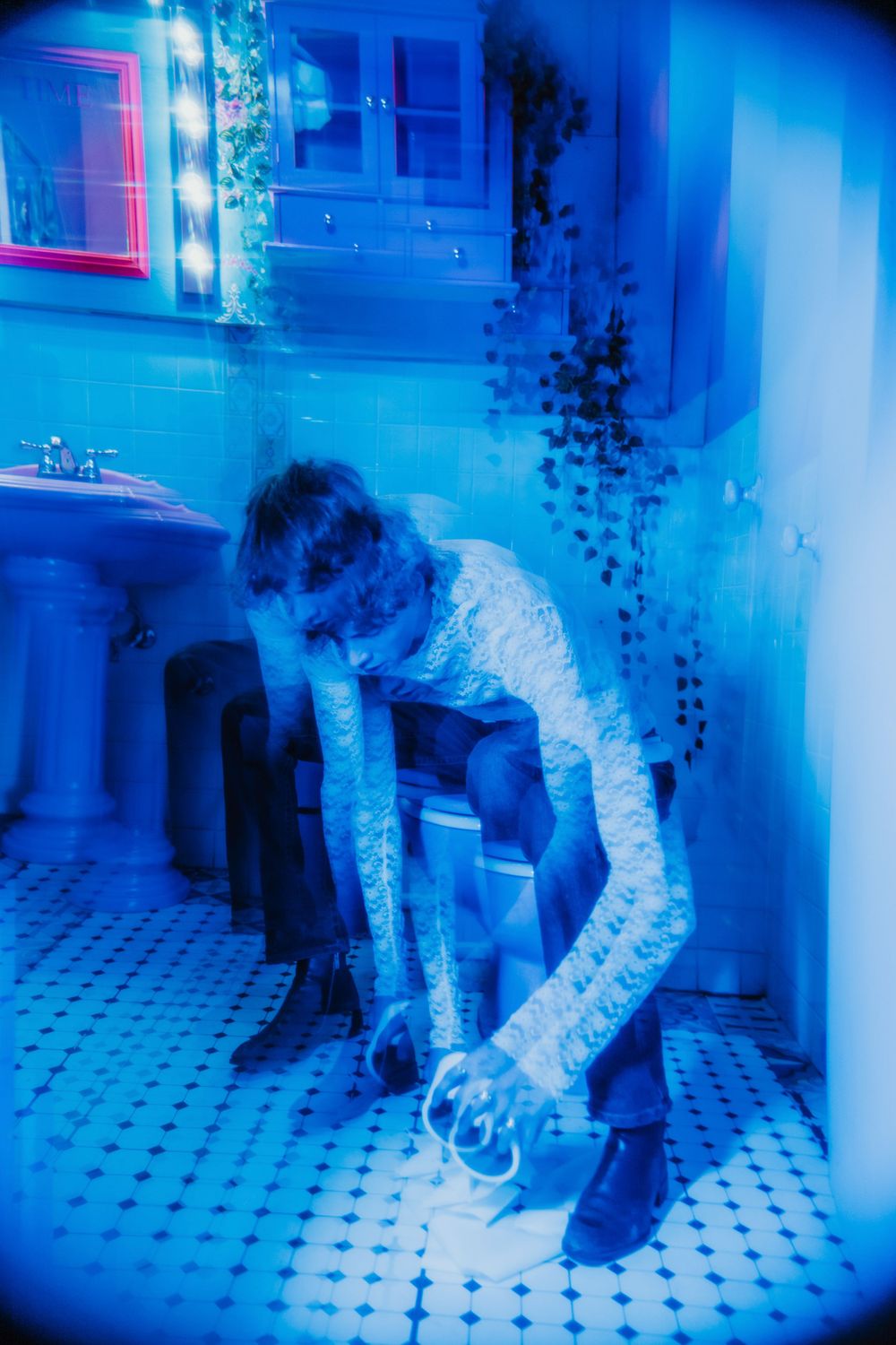 Figure in blue-tinted bathroom leans down to adjust something on tiled floor while bathed in moody neon lighting.