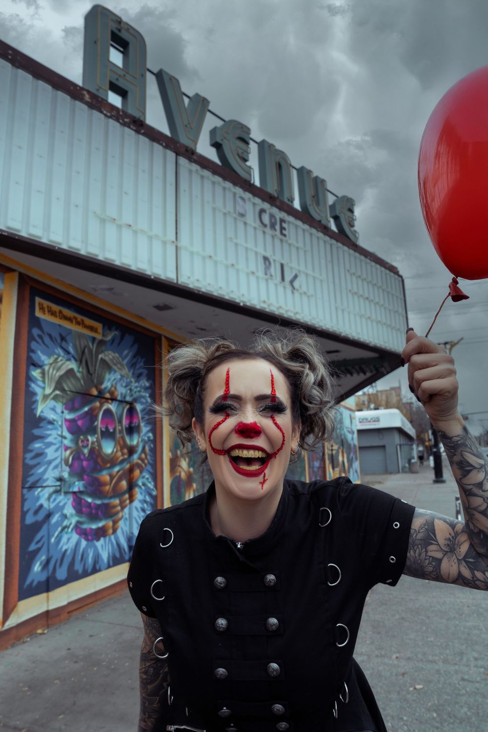 Someone in clown makeup wearing a black jacket holds a red balloon outside a retro movie theater facade.