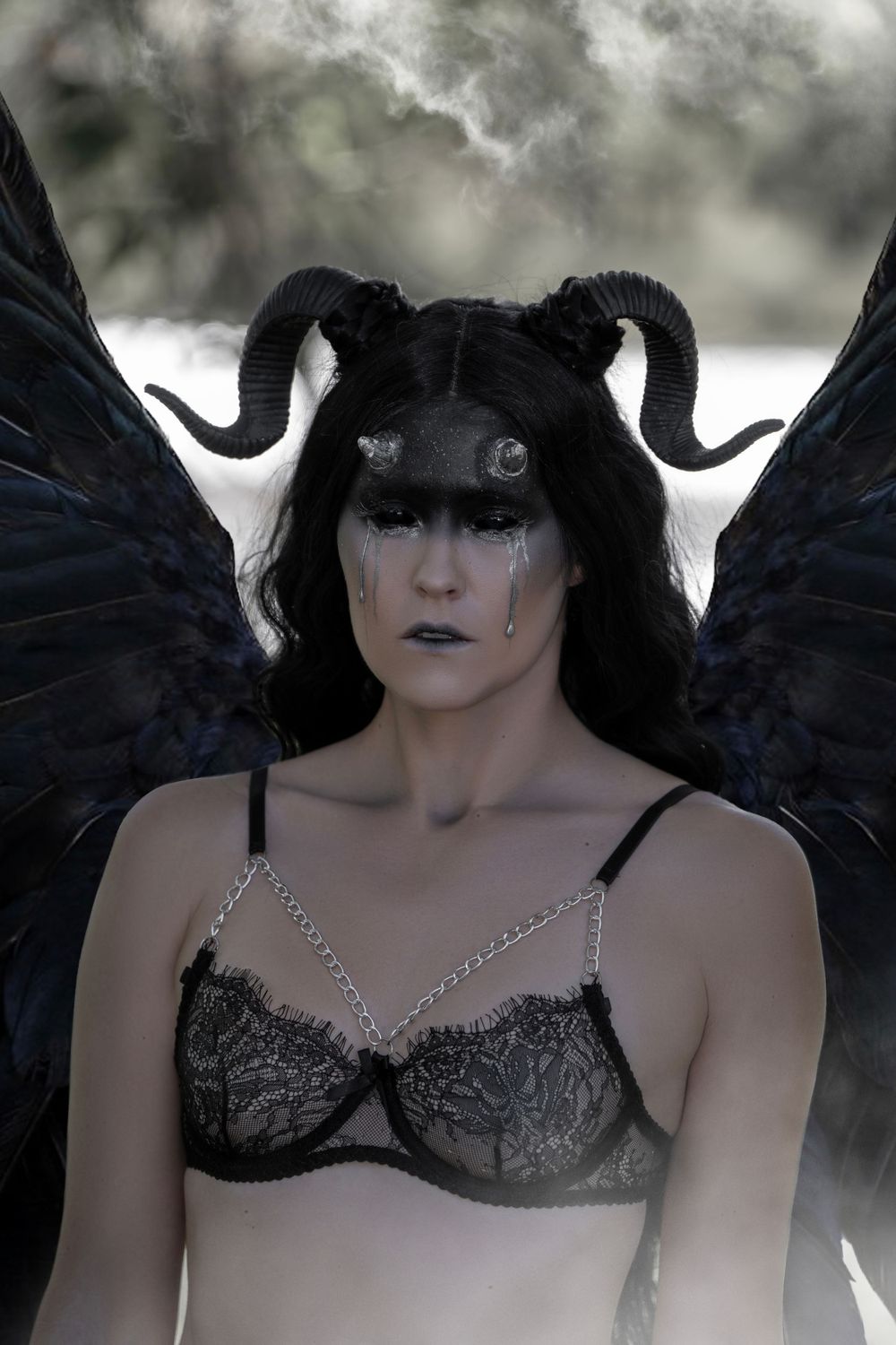 Dark fantasy figure with black horns and wings wearing black lace lingerie against moody cloudy background.