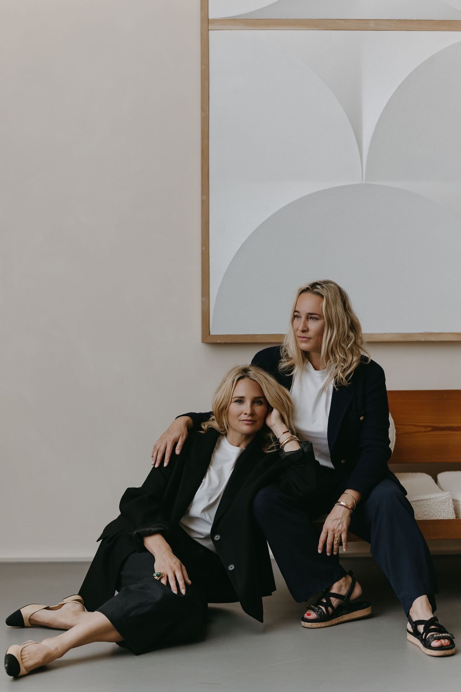 Minimalist portrait of two people in black coats against white wall with geometric artwork.