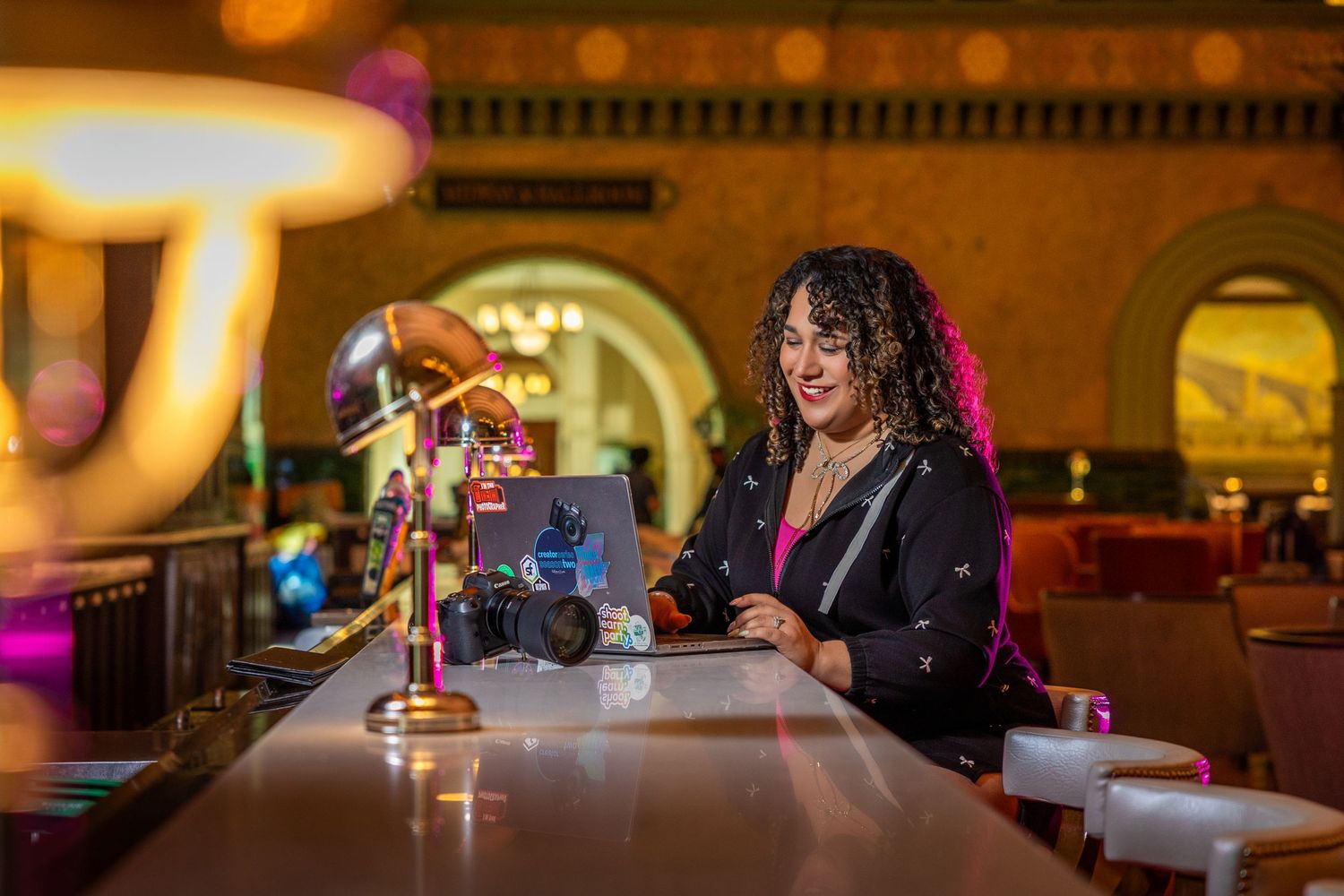 Carolina Waldroff works on her laptop at a glowing bar inside Union Station in St. Louis, camera beside her and vibrant lights surrounding.