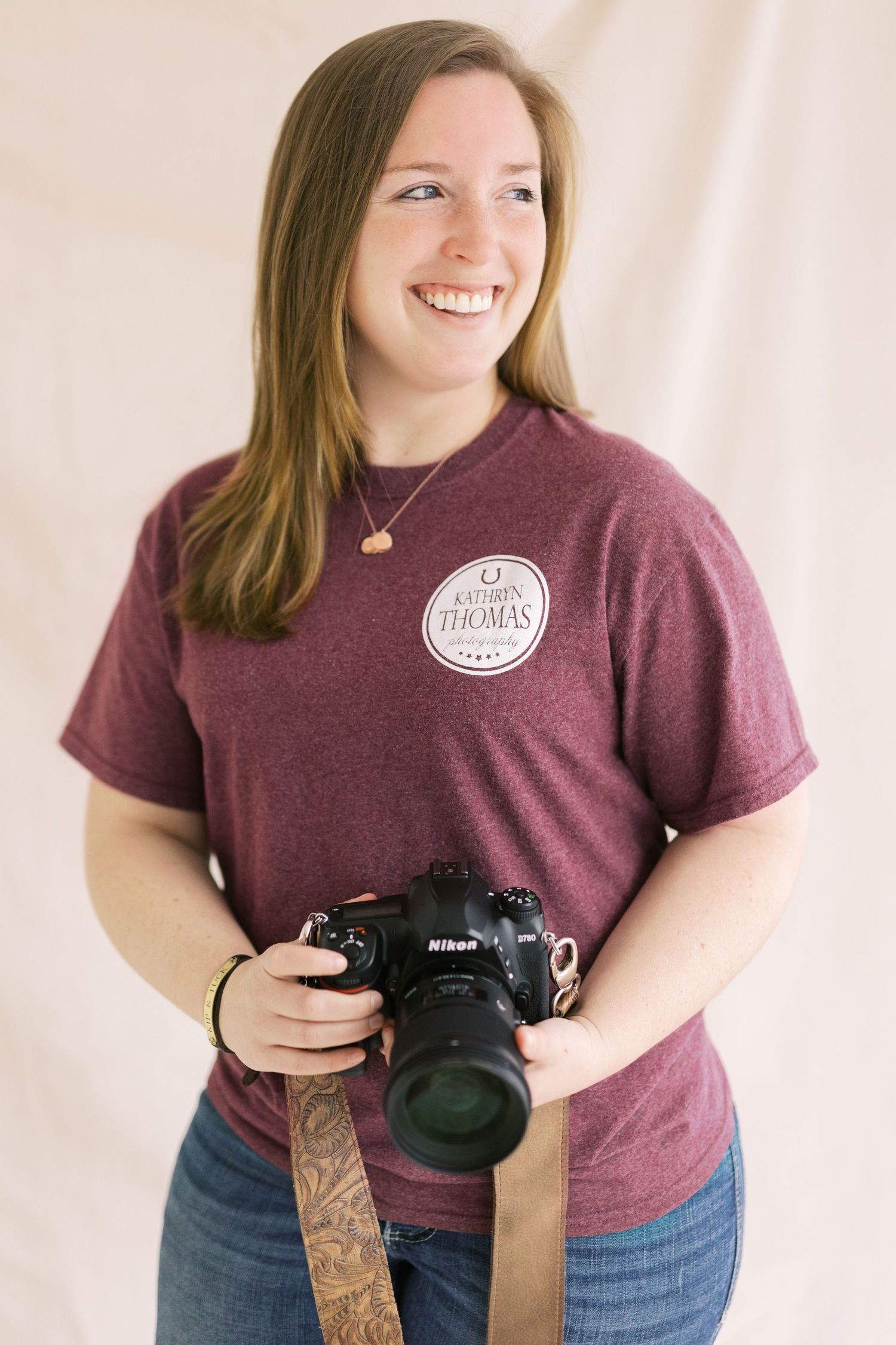 A photographer wearing a burgundy t-shirt holds a professional camera with a lens strap.