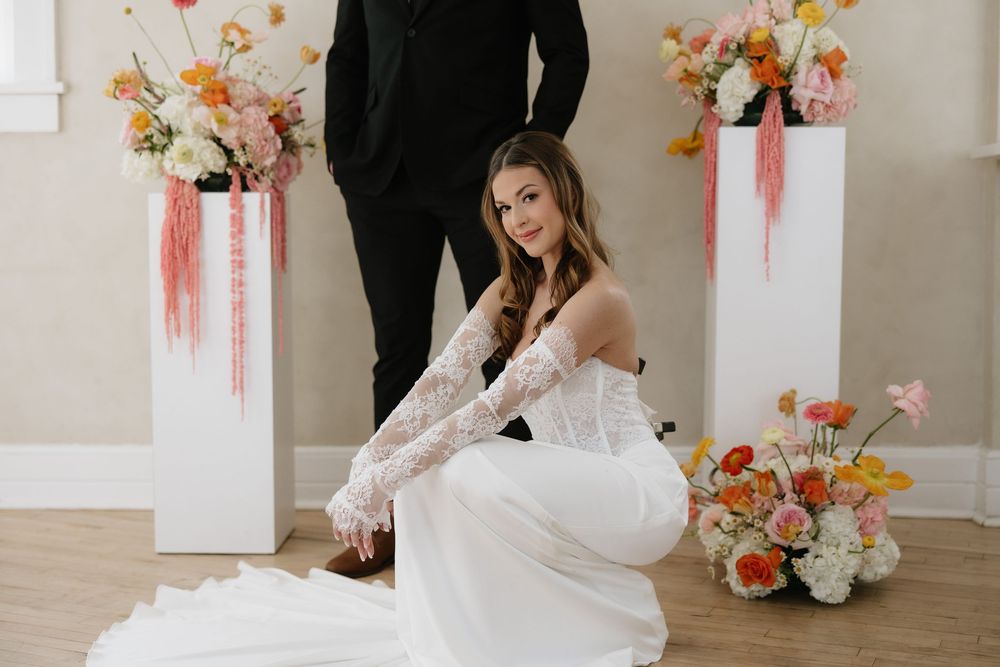 Bride in white gown with lace gloves poses by floral pedestals as groom stands behind her.