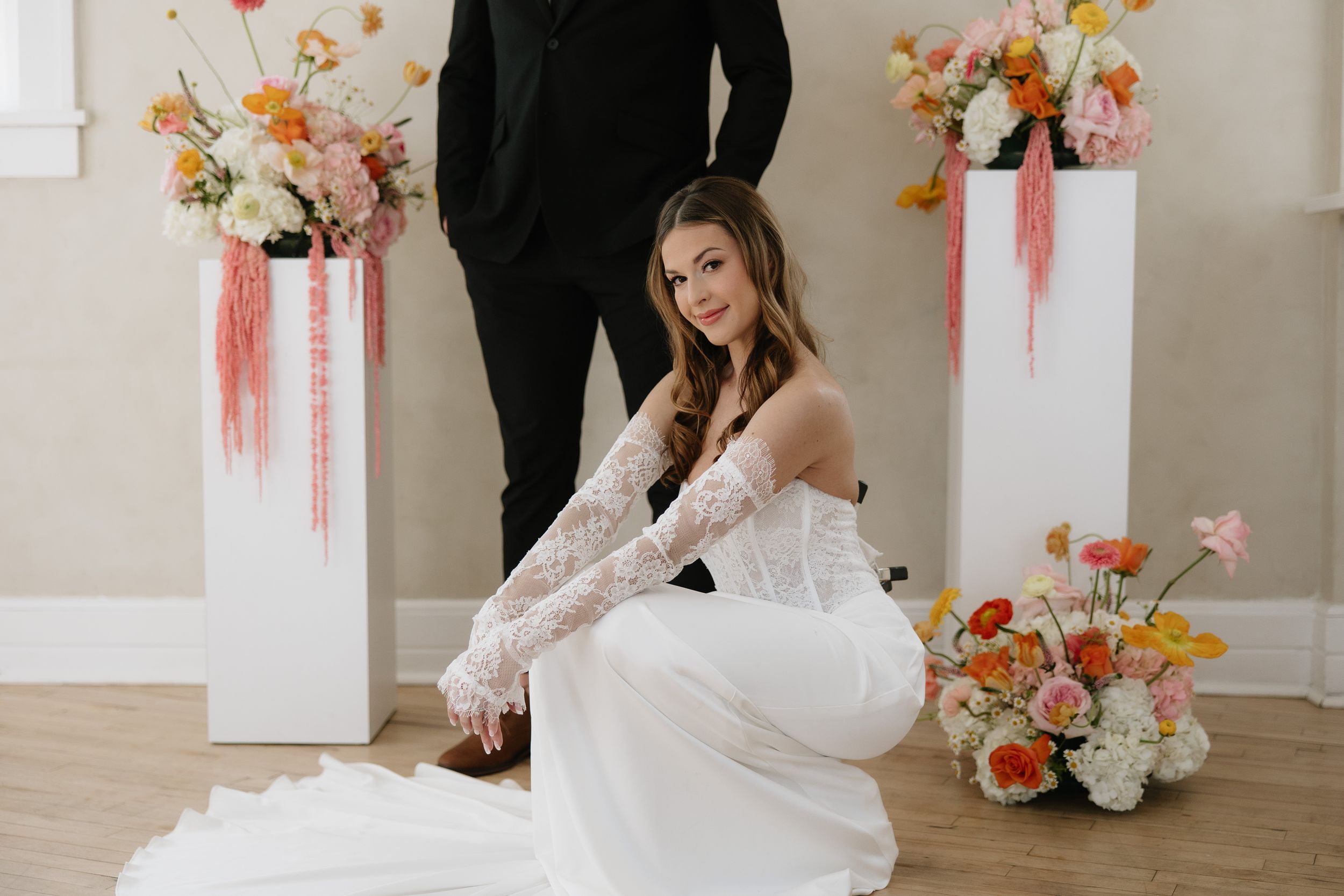 Bride in white gown with lace gloves poses by floral pedestals as groom stands behind her.