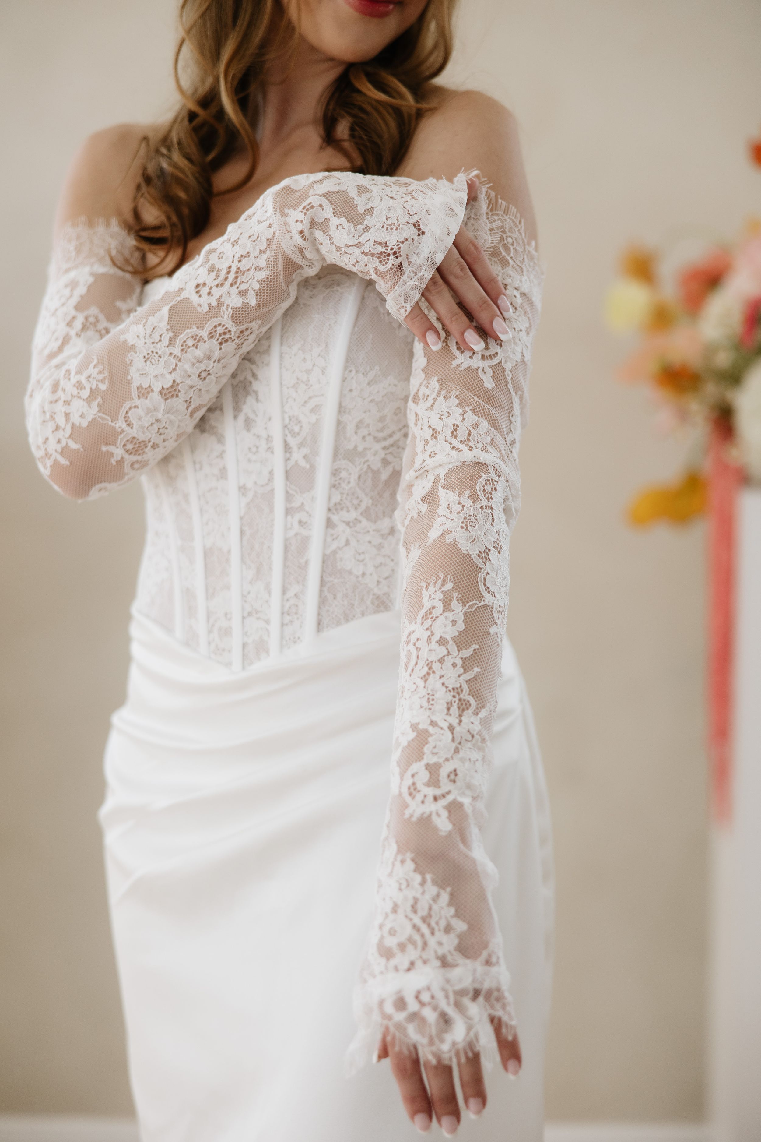 Bride wearing an elegant off-shoulder lace corset wedding gown with sheer long lace gloves, posing gracefully indoors.