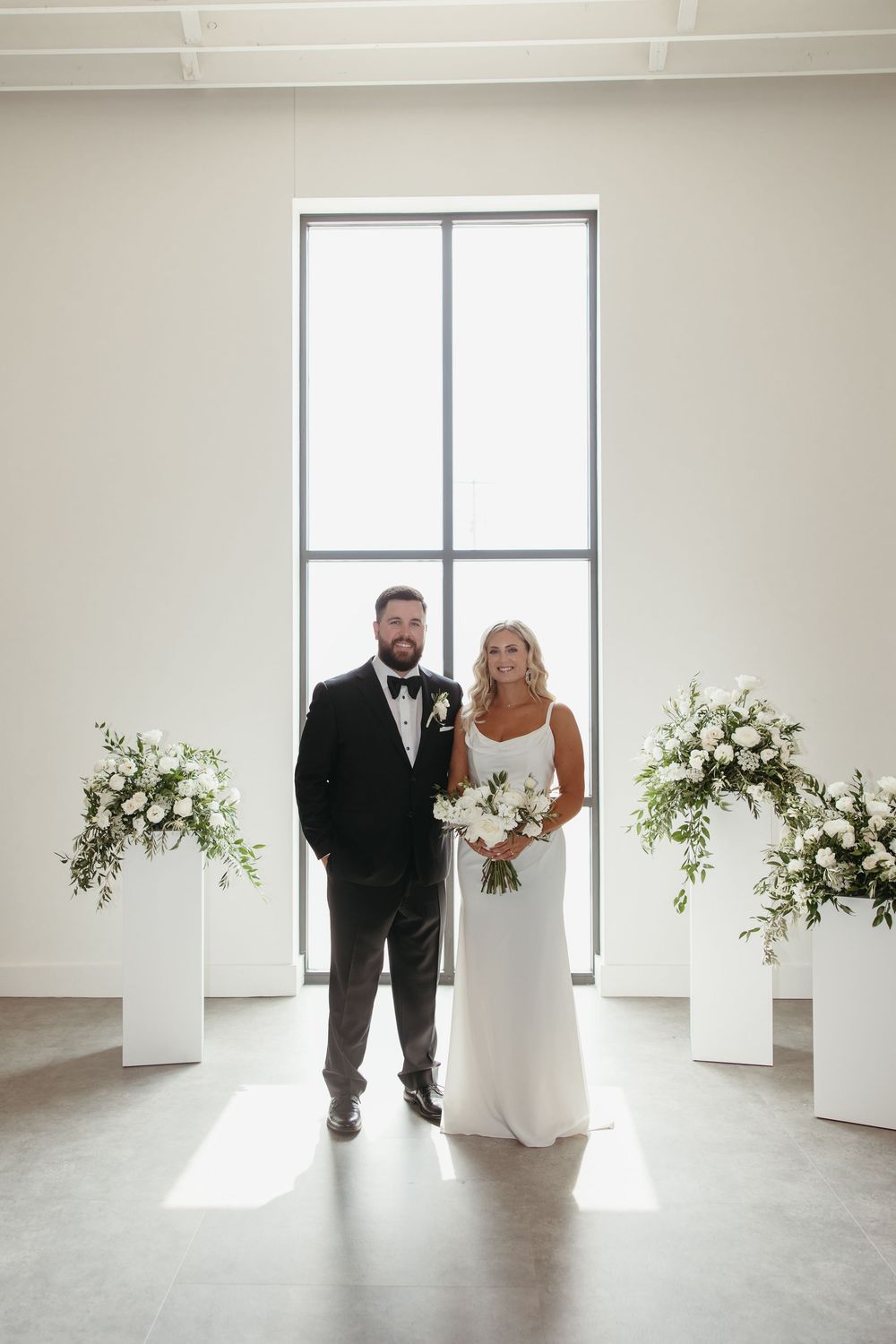 Bride and groom pose together at modern wedding altar with white floral arrangements and large window backdrop.