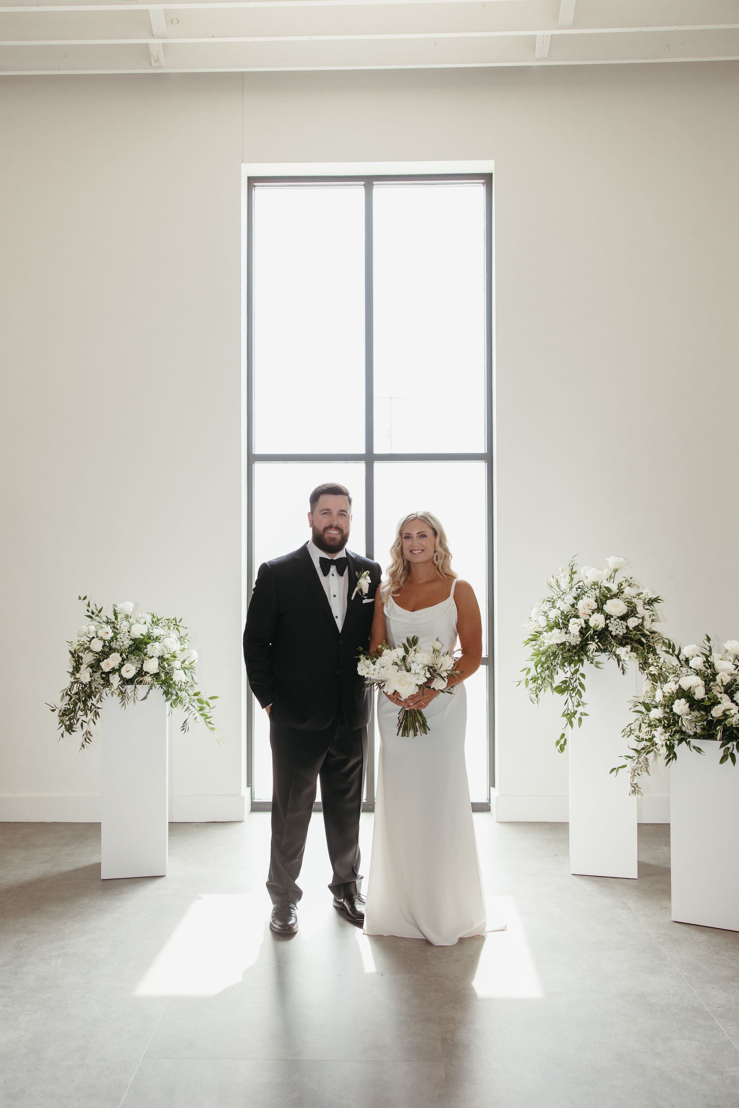 Bride and groom pose together at modern wedding altar with white floral arrangements and large window backdrop.