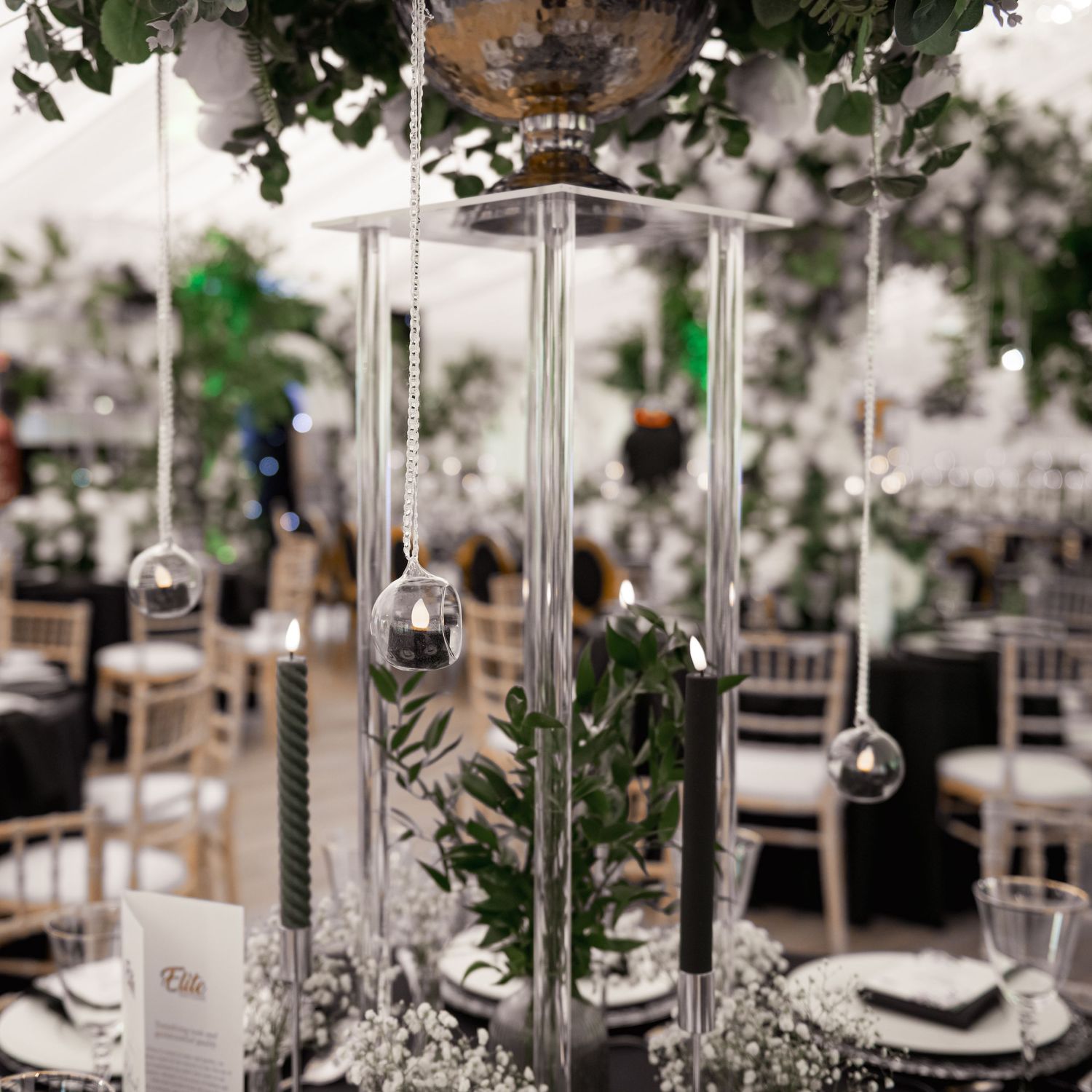 Elegant wedding centerpiece with tall glass lantern, greenery, and hanging silver ornaments on table.