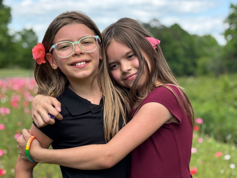 Two people embrace and smile while standing in a lush green field with pink flowers in the background.