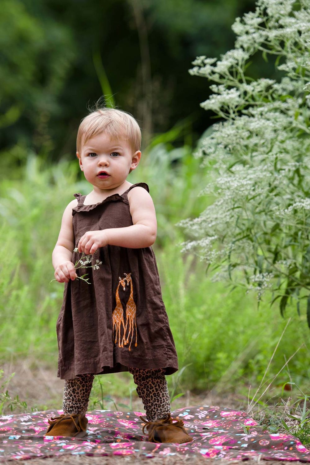 A small child in a brown dress and polka dot leggings stands among blooming white wildflowers in a garden.