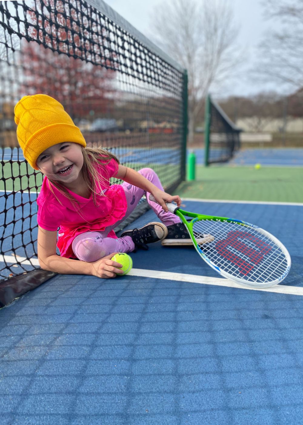 Young tennis player in yellow beanie and pink shirt smiling while posing with racket on court.