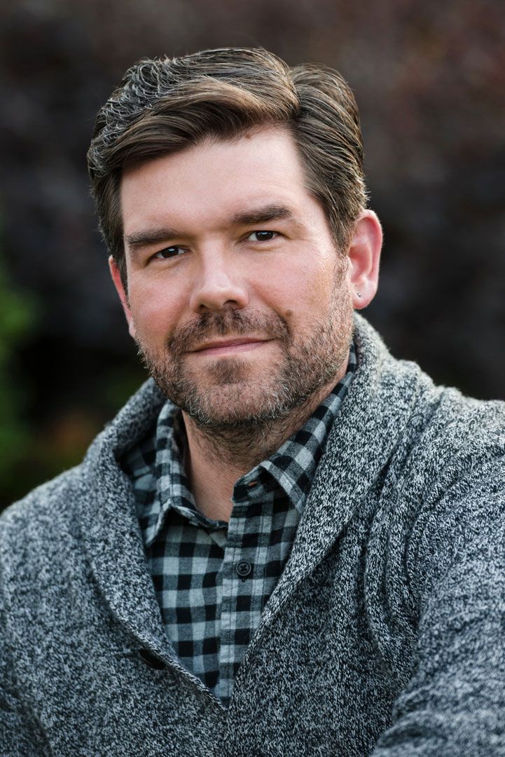 A person wearing a grey cardigan over a black and white checkered shirt poses for a casual outdoor portrait.