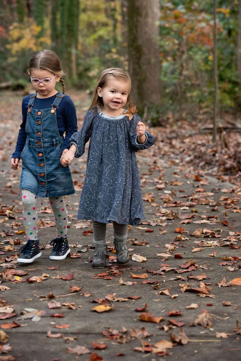 Two young children walking together on a leaf-covered path in autumn forest scenery.