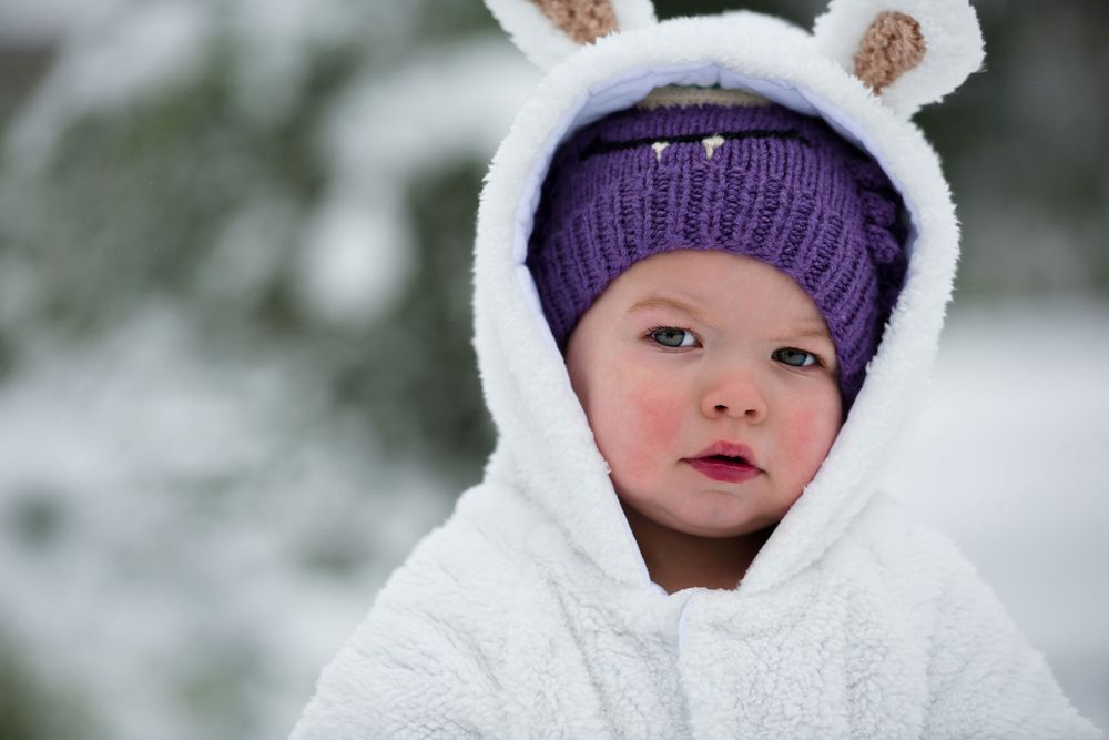Child wearing white hooded winter coat with bunny ears and purple knit hat in snowy setting.