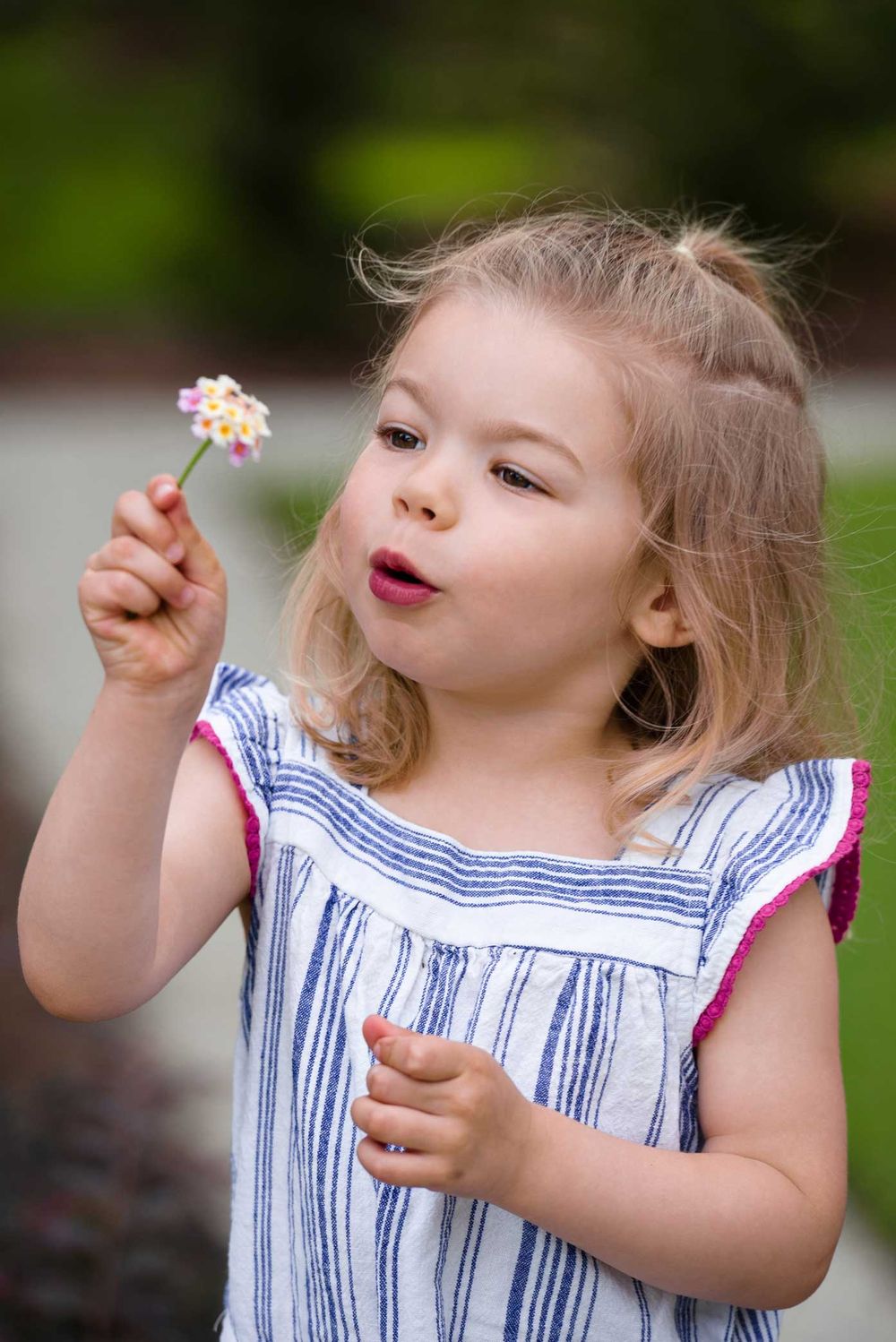Child in a striped dress holding a small flower while making a playful expression in an outdoor setting.